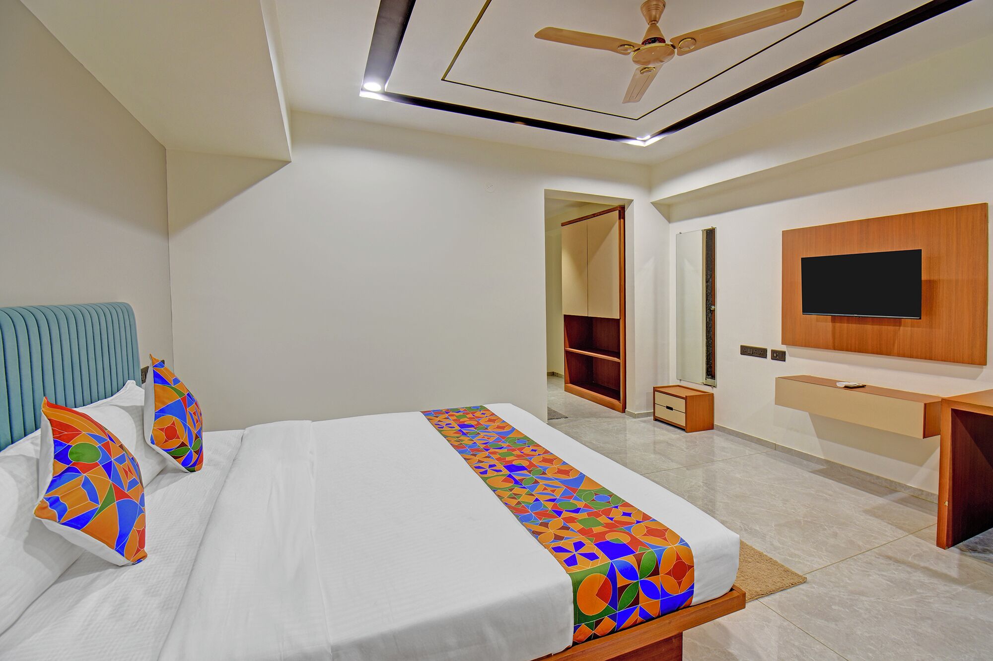 Une chambre d’hôtel moderne équipée d’un lit, d’un ventilateur de plafond, d’une télévision, d’une armoire et d’une salle de bain visible à travers une porte ouverte.