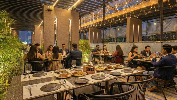 Ένα εστιατόριο με τραπέζια σε εξωτερικό χώρο, στρωμένα με πιάτα, ποτήρια και μαχαιροπίρουνα, όπου οι θαμώνες απολαμβάνουν το γεύμα τους.