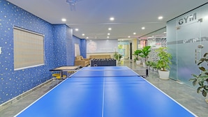 A room with a blue ping pong table, a ceiling fan, and a large window with blinds. - Fabhotel Aashray (Hyderabad)