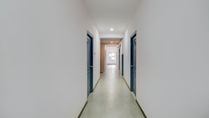 A long, narrow hallway with white walls and blue doors, leading to a room with a window and a door at the end. - Fabhotel Rk Grand (Hyderabad)