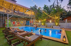 Área de piscina com espreguiçadeiras, um espaço de estar coberto e um mural.