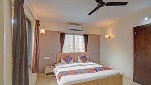 A bedroom with a bed, a ceiling fan, an air conditioning unit, a nightstand, and a window with curtains. - Fabhotel Panchsheel Crown (Pune)