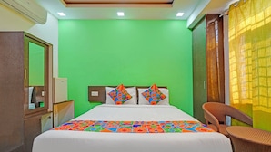 Une chambre d’hôtel avec un mur vert, un lit recouvert d’une couvre-lit aux motifs géométriques colorés, deux oreillers aux motifs géométriques, une chaise en osier et un miroir sur le mur de gauche.