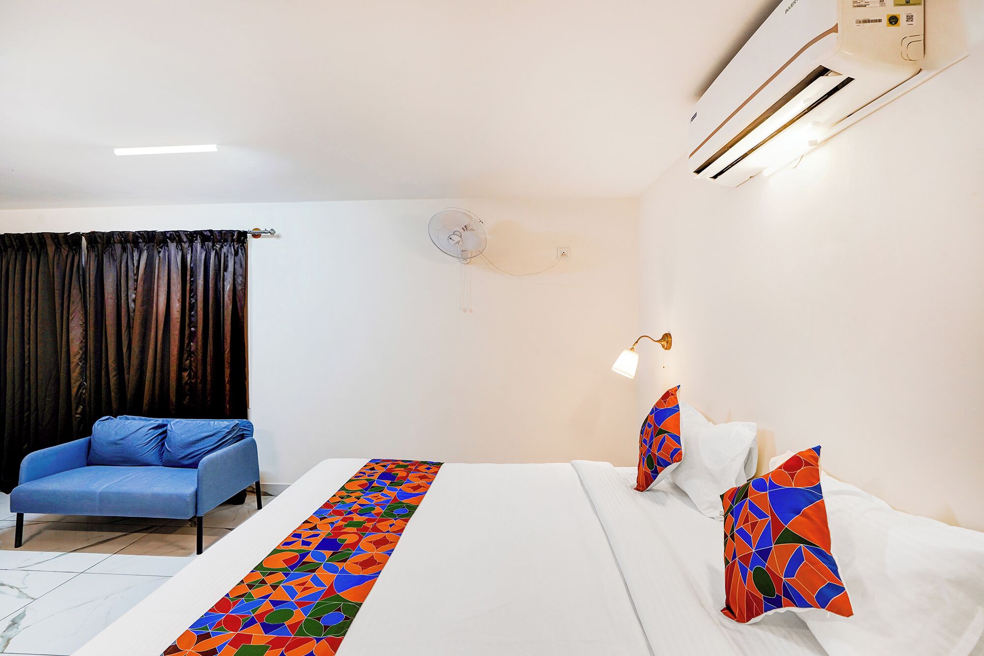 Una habitación de hotel con una cama, un sofá azul, un ventilador de pared y un cojín decorativo.
