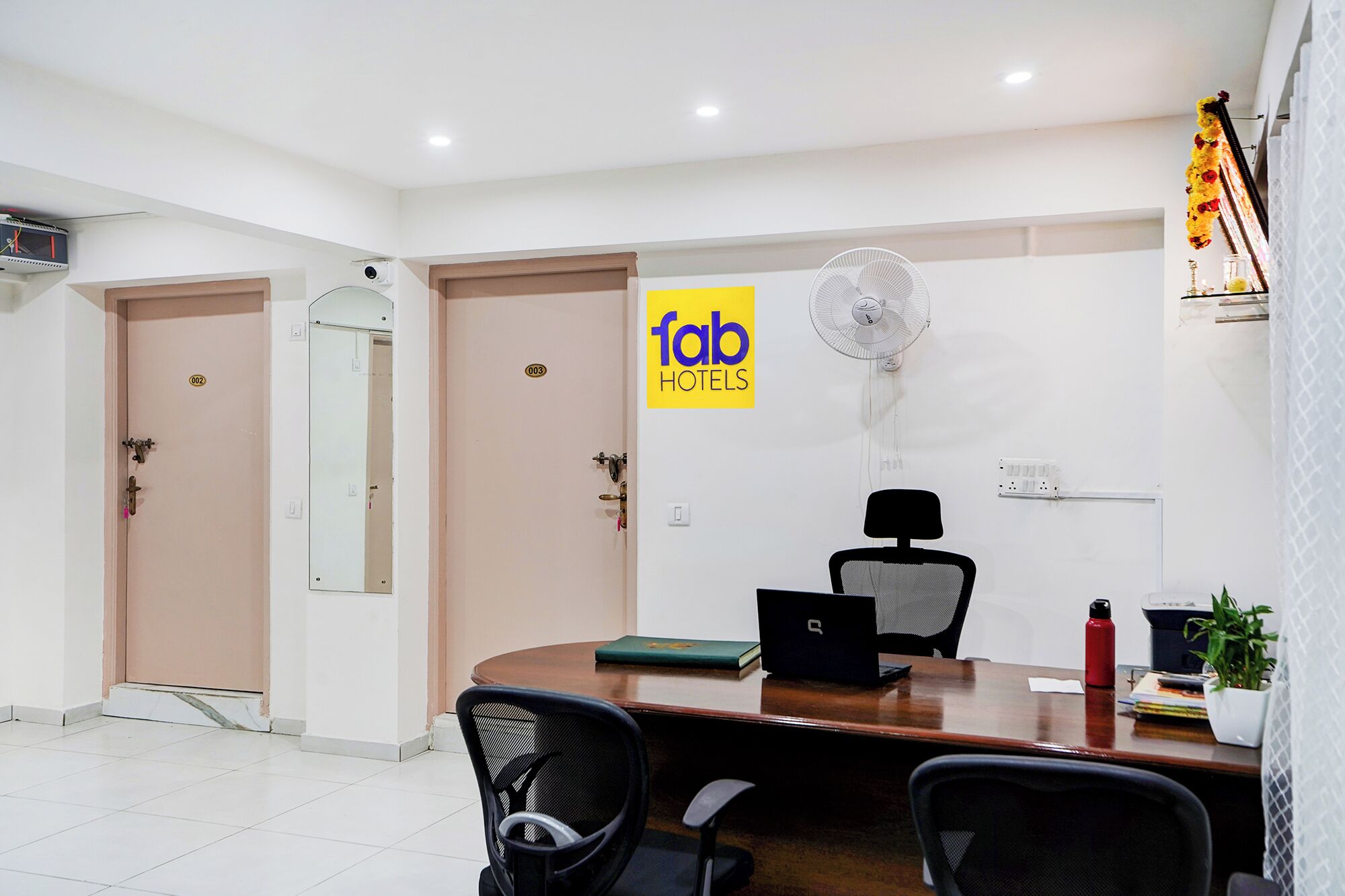 A conference room with a wooden table, black office chairs, a laptop, a desktop computer, a standing fan, and a 'fab hotels' sign on the wall.