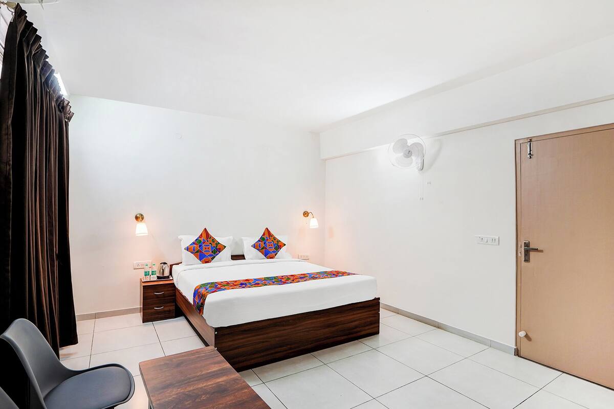 A hotel room with a bed, bedside tables, a desk, a chair, a wardrobe, and a wall-mounted fan.