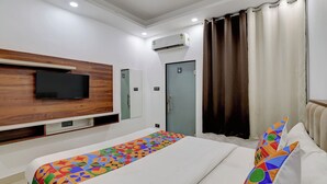 A modern hotel room with a large bed, a flat-screen TV mounted on a wooden panel, a built-in air conditioning unit, and a window with curtains. - Fabhotel Groom Room (Noida)
