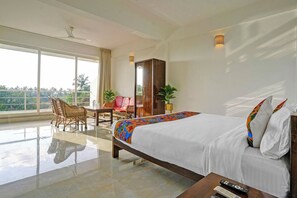 Ein Hotelzimmer mit einem großen Bett, einer Sitzecke mit Stühlen, einem Holzschrank und Blick auf Palmen durch große Fenster.