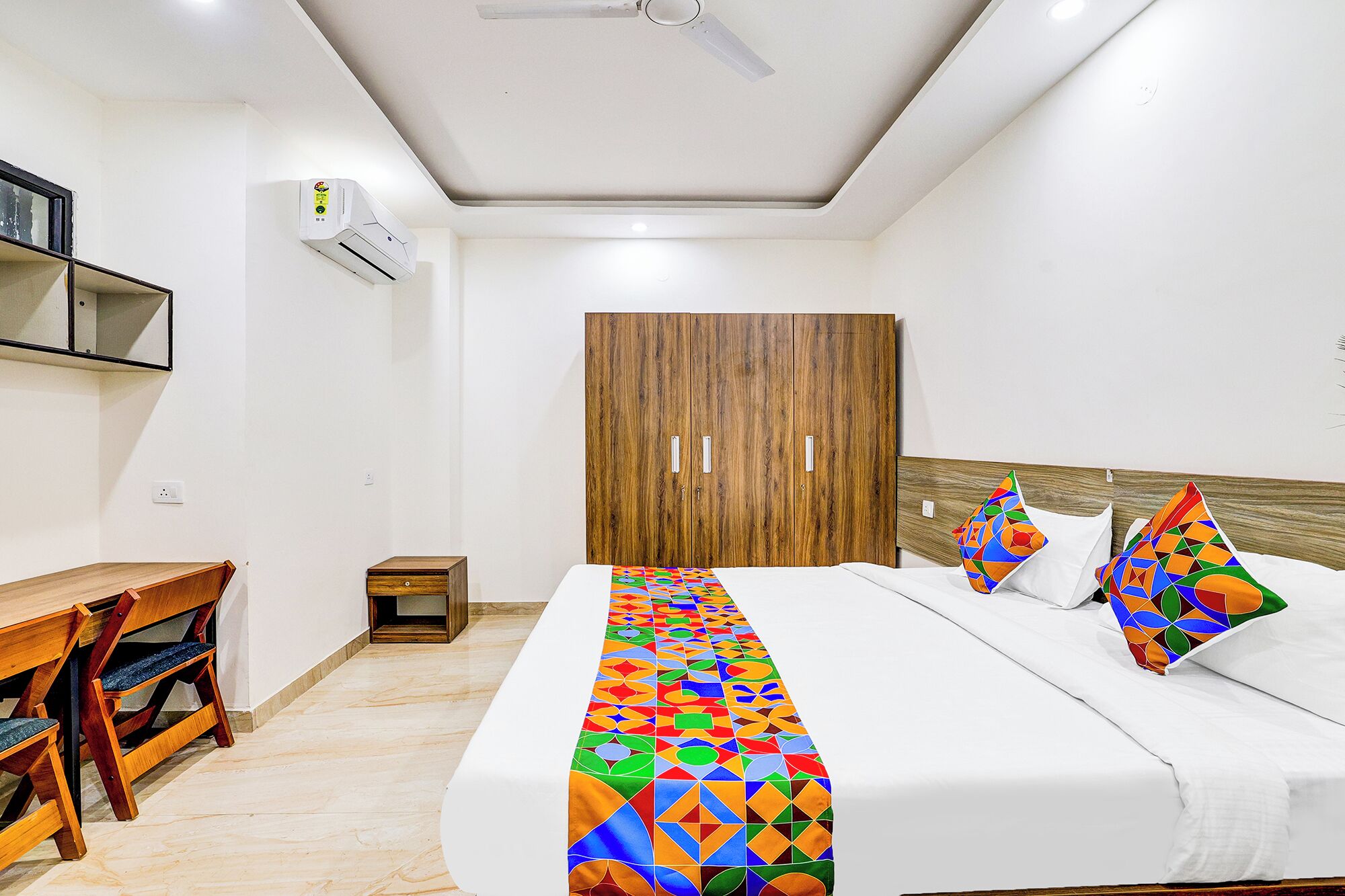A bedroom with a bed, a wooden wardrobe, a desk with chairs, and a ceiling fan.