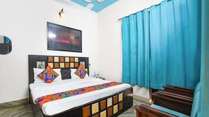 A hotel room with a bed, a headboard with a geometric pattern, a framed artwork above the bed, a blue curtain, and a wooden bench. - Fabhotel Decent (New Delhi)