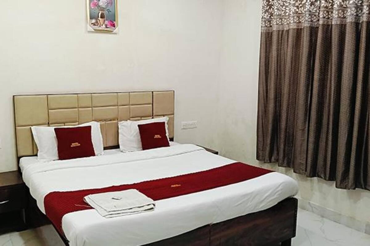 A hotel room with a bed, two pillows, a nightstand, a red throw blanket, a window with curtains, and a wall-mounted picture.