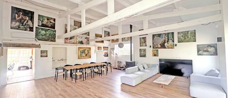 A spacious room with a high ceiling, wooden flooring, a long dining table, and multiple seating arrangements.