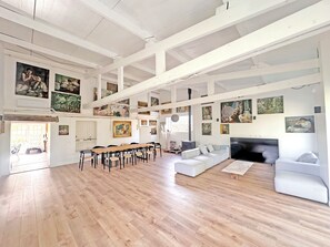 A spacious room with a high ceiling, wooden flooring, a long dining table, and multiple seating arrangements. - L'Atelier, between the beach and nature (Barneville-Carteret)
