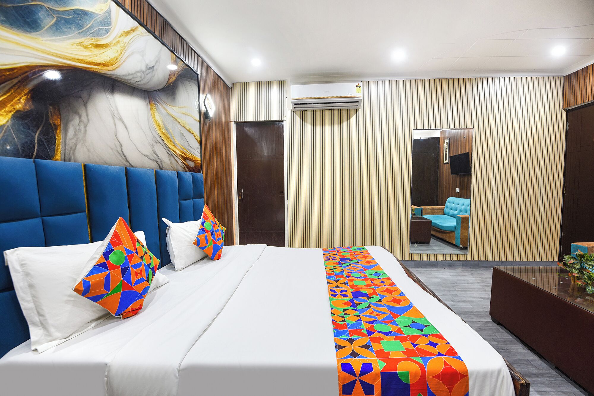A modern hotel room with a large bed, a blue headboard, a colorful geometric bedspread, and a large abstract painting on the wall.