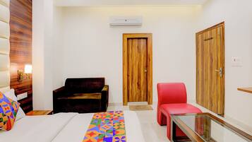 A hotel room with a bed, a red chair, a wooden wardrobe, and a small table with a lamp.
