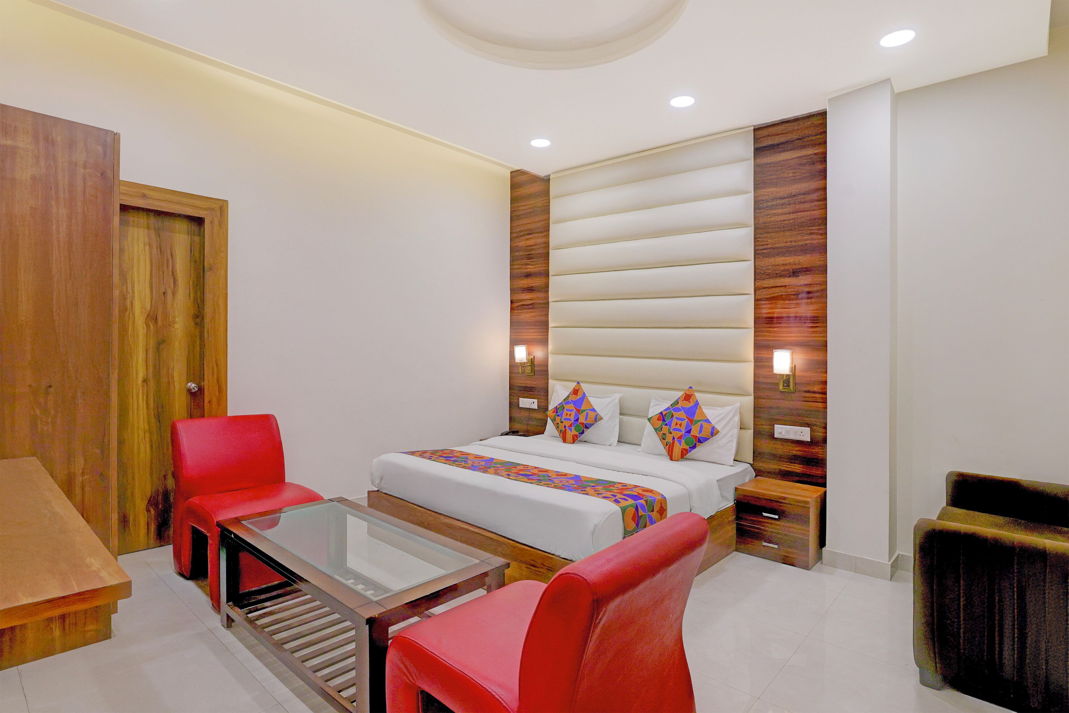 Una habitación de hotel con una cama, dos sillas rojas, una mesa con superficie de vidrio, muebles de madera y una puerta de madera.