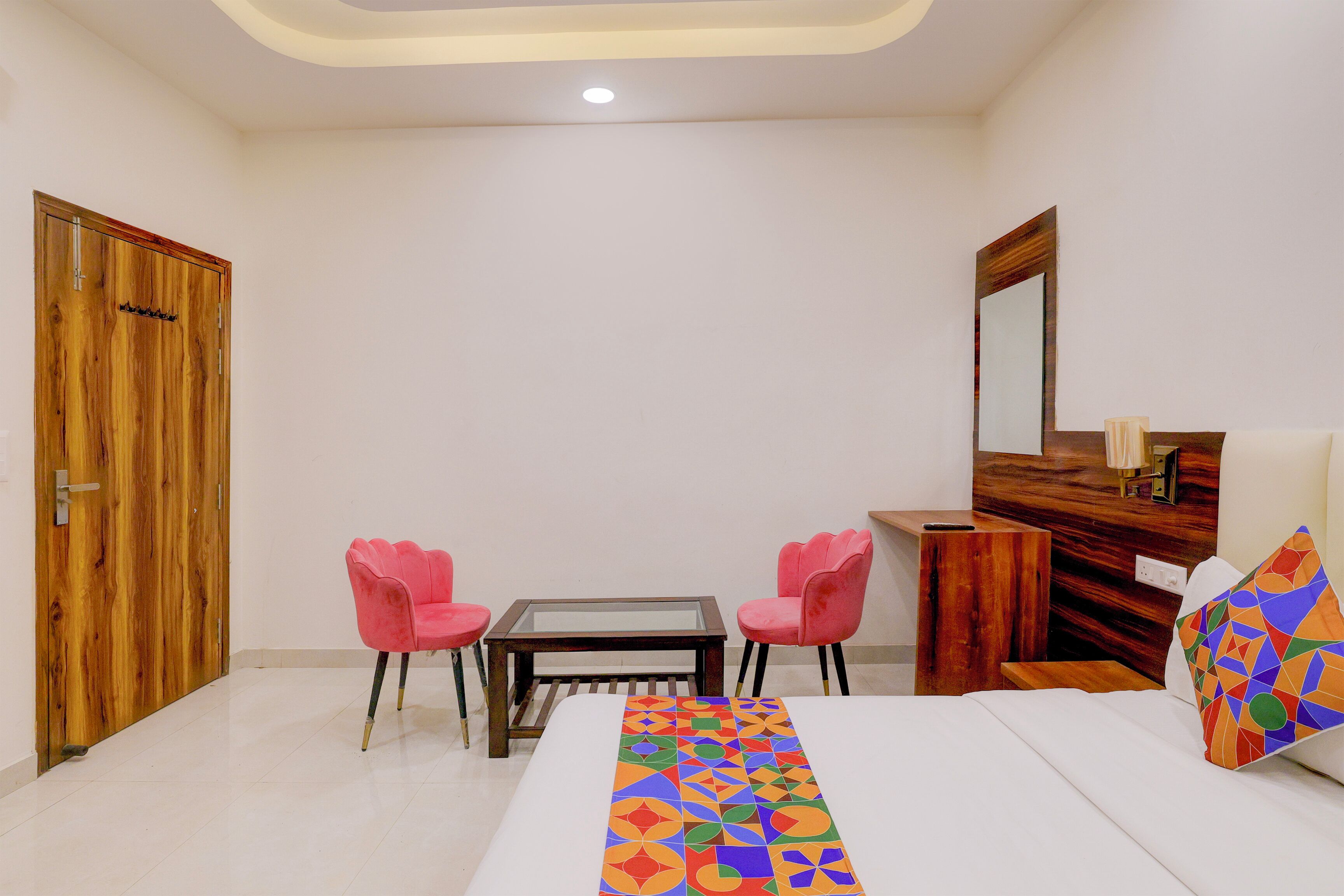 A hotel room with a bed, a wooden wardrobe, a small table with two pink chairs, and a mirror on the wall.