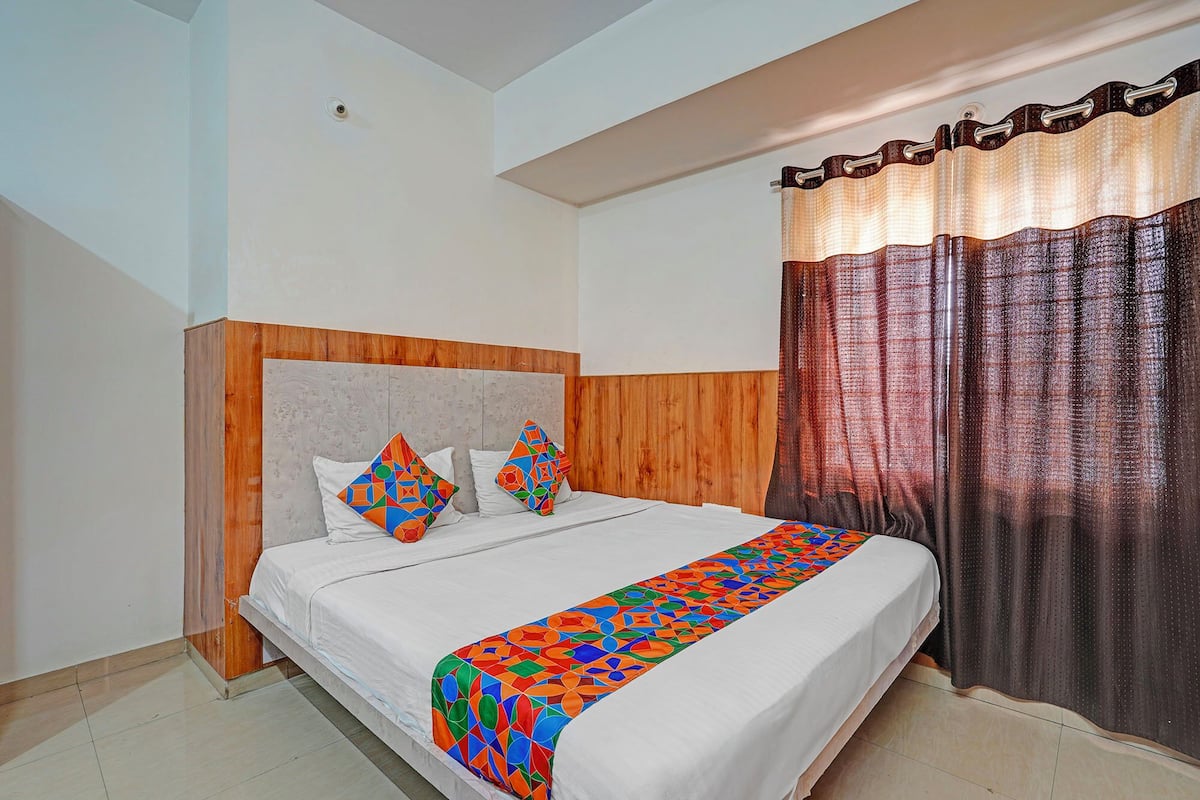 A bedroom with a bed, wooden headboard, two pillows, a colorful bedspread, and a curtain.