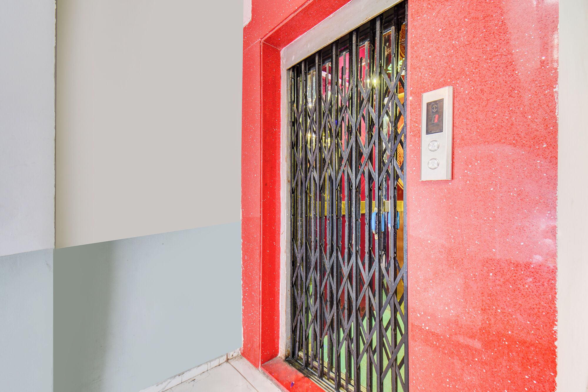 Una puerta con una reja de seguridad metálica, una pared con una decoración roja y blanca, y un pequeño panel de control en la pared.