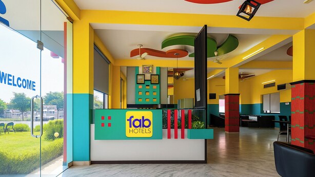 Un mostrador de recepción de hotel con un cartel de 'fab hotels', una pared de cristal con un letrero de 'BIENVENIDOS' y vistas a zonas verdes.