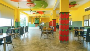 Un comedor con techos altos, diseños coloridos en el techo y un columnal central con un motivo rojo y verde. Mesas y sillas dispuestas para comer.