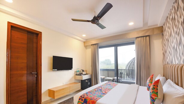 A hotel room with a bed, a TV, a ceiling fan, a window with curtains, and a balcony with a chair. - FabHotel Hygge Stay (Gurugram)