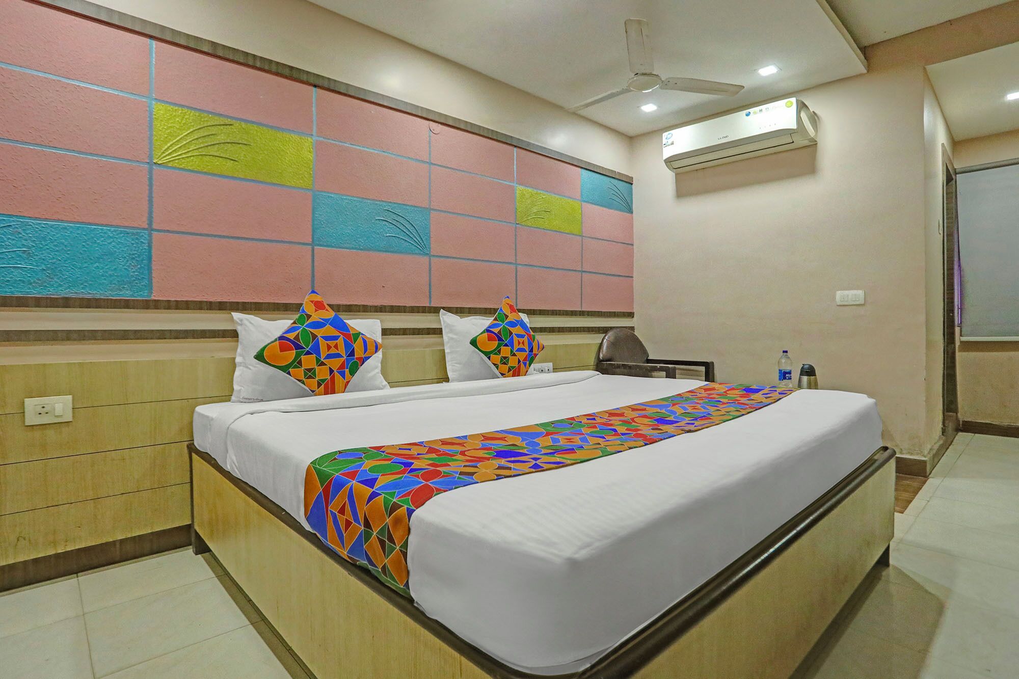 A hotel room with a bed, a desk, an air conditioning unit, and a wall with a colorful geometric pattern.