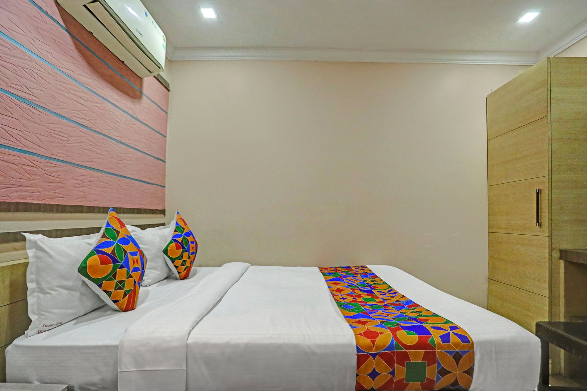 Habitación de hotel con cama, ropa de cama blanca, almohadas de colores, un plaid estampado y un armario de madera.