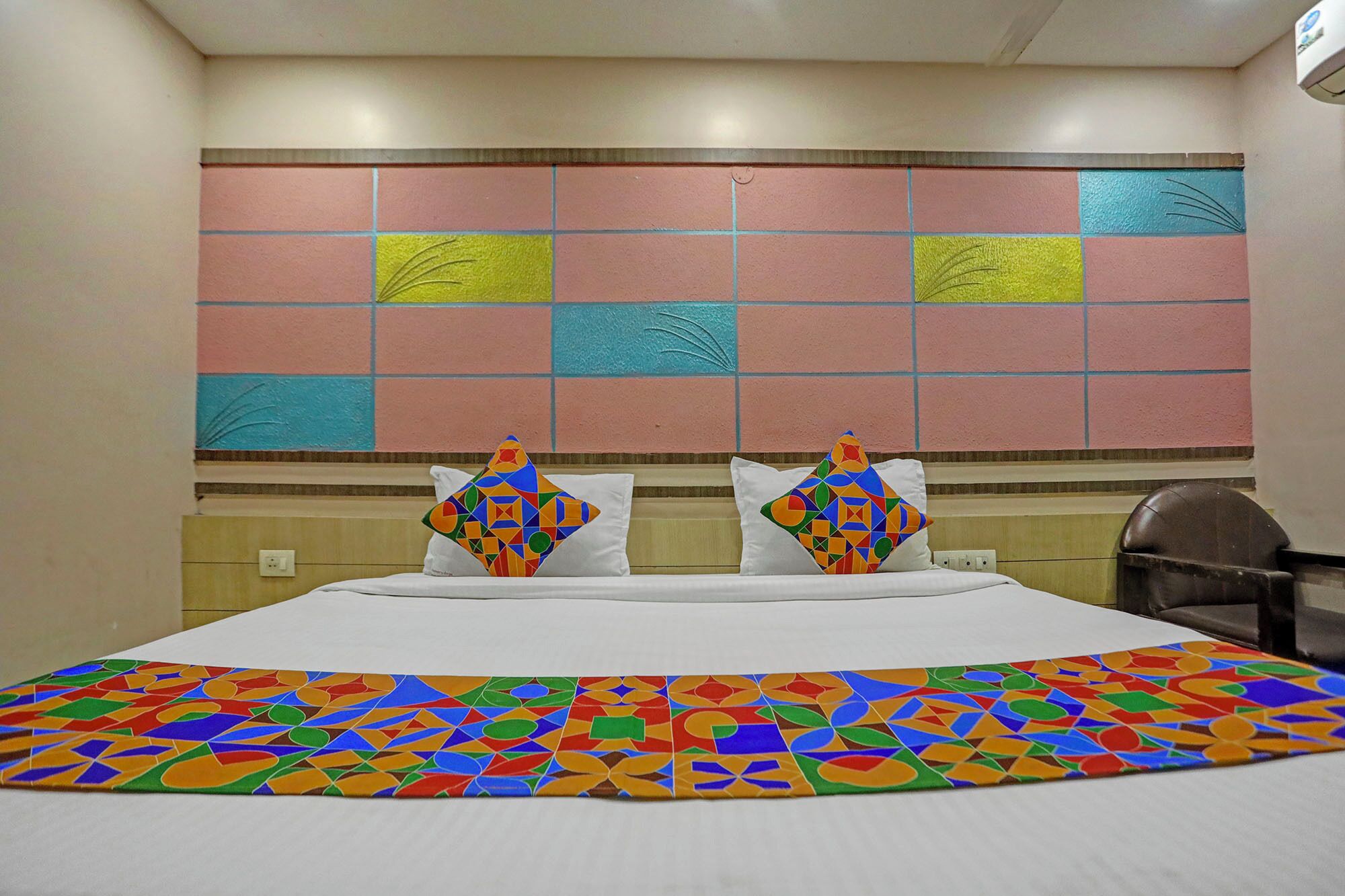 Quarto de hotel com uma cama grande, almofadas coloridas com padrão geométrico, um tapete vibrante e uma parede decorativa com padrão de grade.