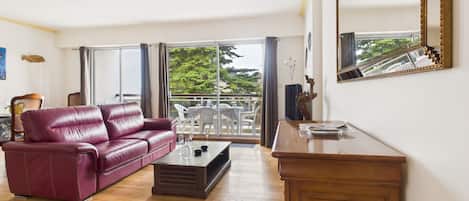 A living room with a leather sofa, a wooden coffee table, and a wooden cabinet. There is a balcony with outdoor furniture and a view of trees.