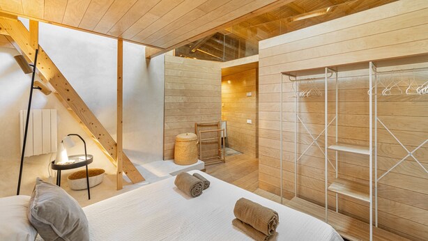A modern hotel room with a wooden ceiling, a bed with white linens, a small bedside table with a lamp, and a wooden wall unit with shelves and a hanging rack.