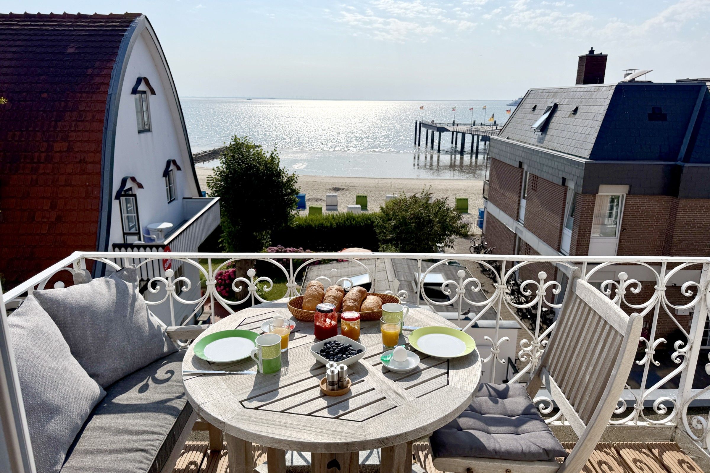 A balcony with a table set for breakfast overlooking a beach and a pier.