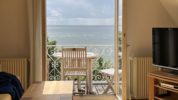 Un balcón con vista al mar, una silla de madera, una mesa y un televisor sobre un soporte.