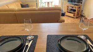 Una mesa de comedor dispuesta con platos, cubiertos y copas de vino, con un sofá y televisor al fondo.
