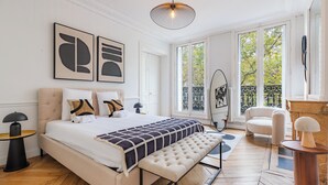 A bedroom with a large bed, a bench, a round table, and a chair. There are two windows with a view of trees. - Appartements 4 boulevard Beaumarchais (Paris)