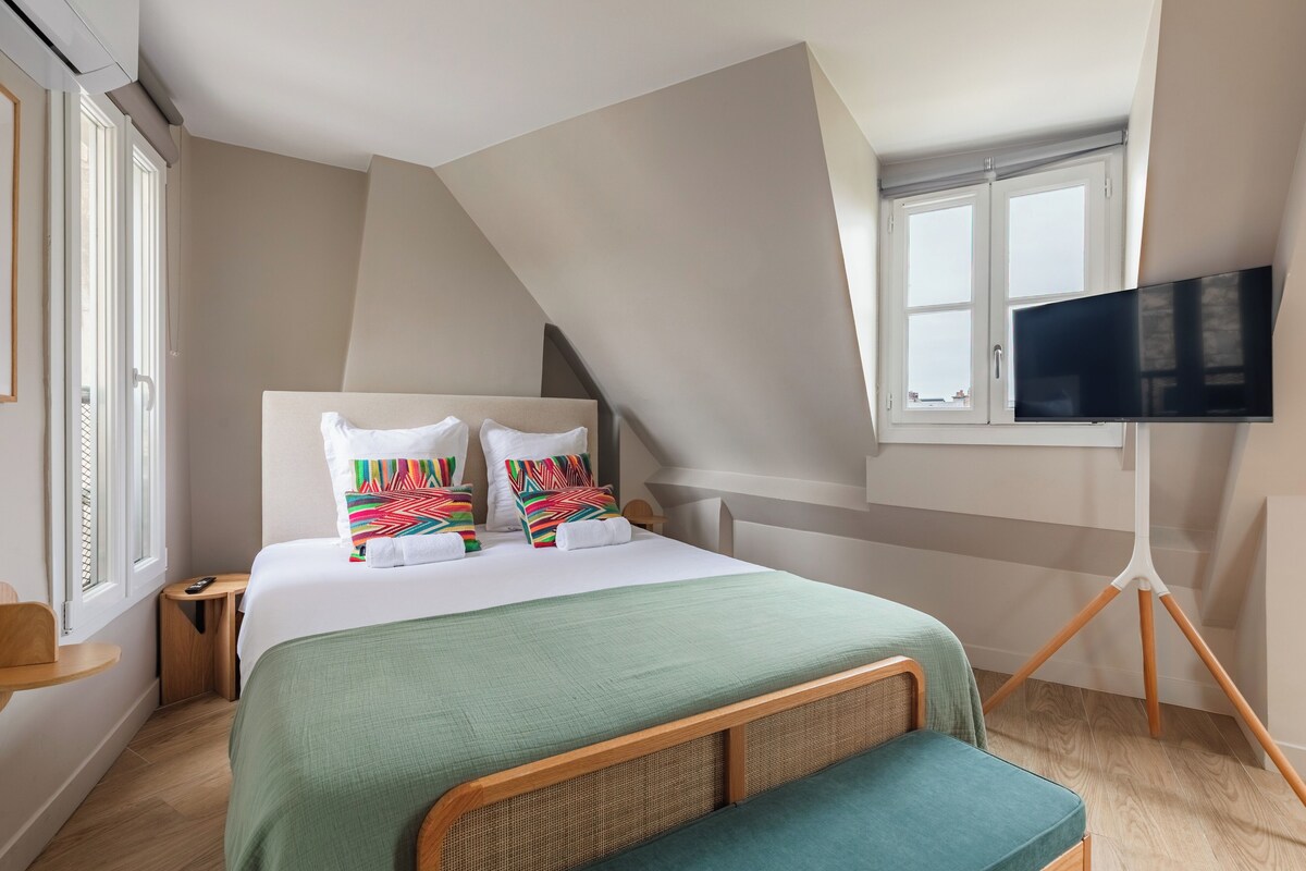 Une chambre moderne avec un grand lit, un téléviseur posé sur un trépied et une fenêtre équipée de stores.