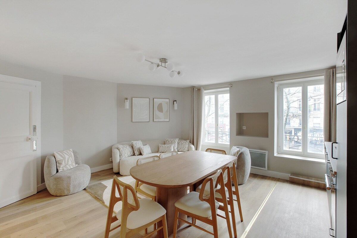 Un salon moderne avec une table à manger en bois, des canapés de couleur claire et une grande fenêtre laissant entrer la lumière naturelle.