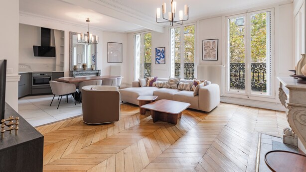 A spacious living room with a herringbone floor, a large sectional sofa, a dining area with a table and chairs, and a kitchen in the background. - Appartements 4 boulevard Beaumarchais (Paris)