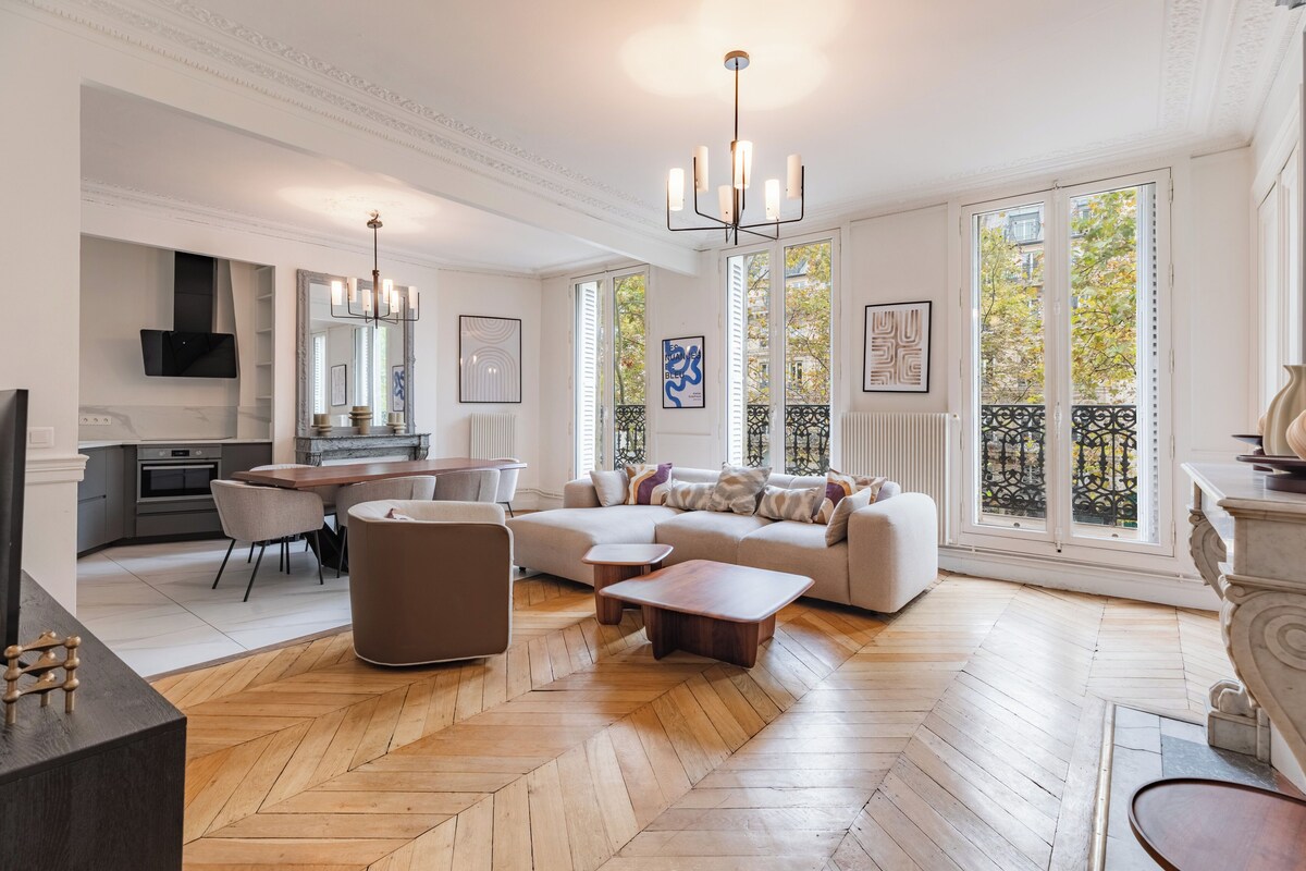 Un vaste salon doté d’un parquet en chevrons, d’un grand canapé d’angle, d’un coin repas avec une table et des chaises, et d’une cuisine en arrière-plan.