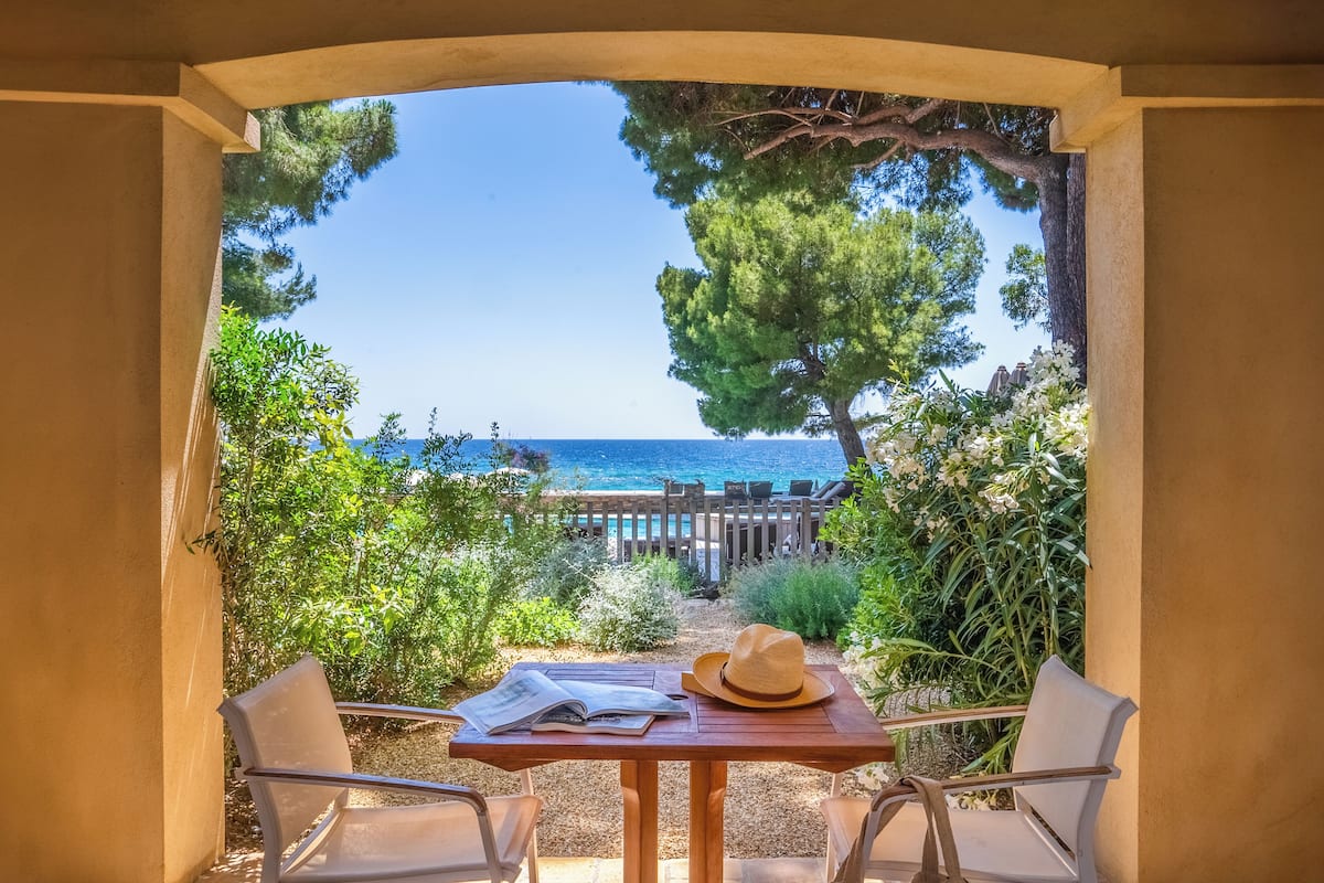 Un patio con una mesa y sillas, un sombrero y un libro, con vistas a una playa con un cerco de madera y árboles.