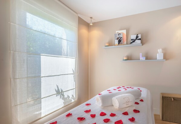 Un cuarto de spa con una cama cubierta de pétalos de rosa, una ventana con cortinas translúcidas y dos estantes con fotografías enmarcadas y velas.