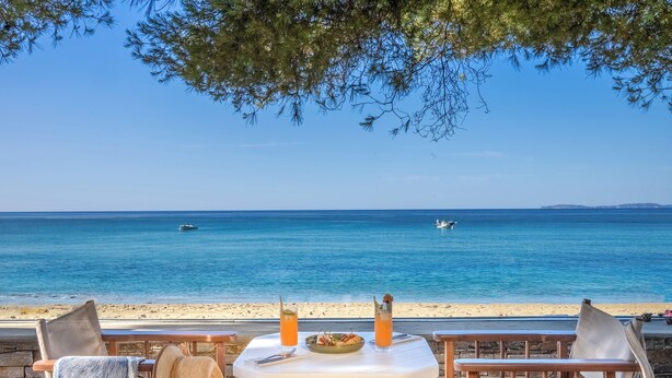 Une table avec une assiette de nourriture et deux boissons, donnant sur une plage avec une eau bleu clair et un bateau au loin.