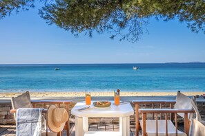 Una mesa con un plato de comida y dos bebidas, con vistas a una playa de aguas azules y un barco en la distancia.