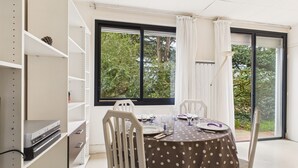 Ein Essbereich mit einem runden Tisch, weißen Stühlen und einer Tischdecke mit Punkten. Ein Bücherregal mit DVD-Player steht daneben, und ein Fenster mit Vorhängen ist ebenfalls zu sehen.