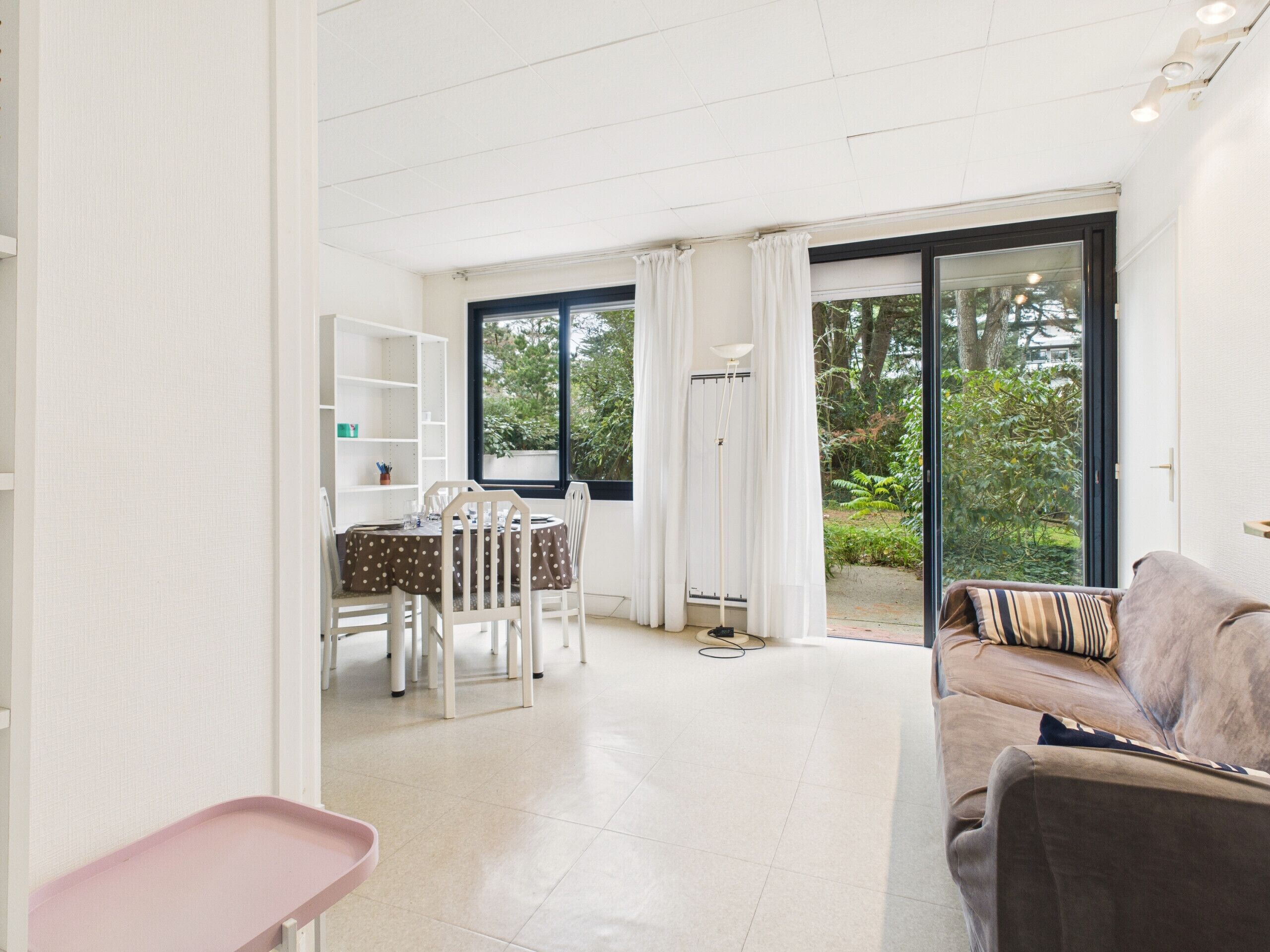 A modern living room with a sofa, a dining area with a table and chairs, and a large sliding glass door leading to a garden.