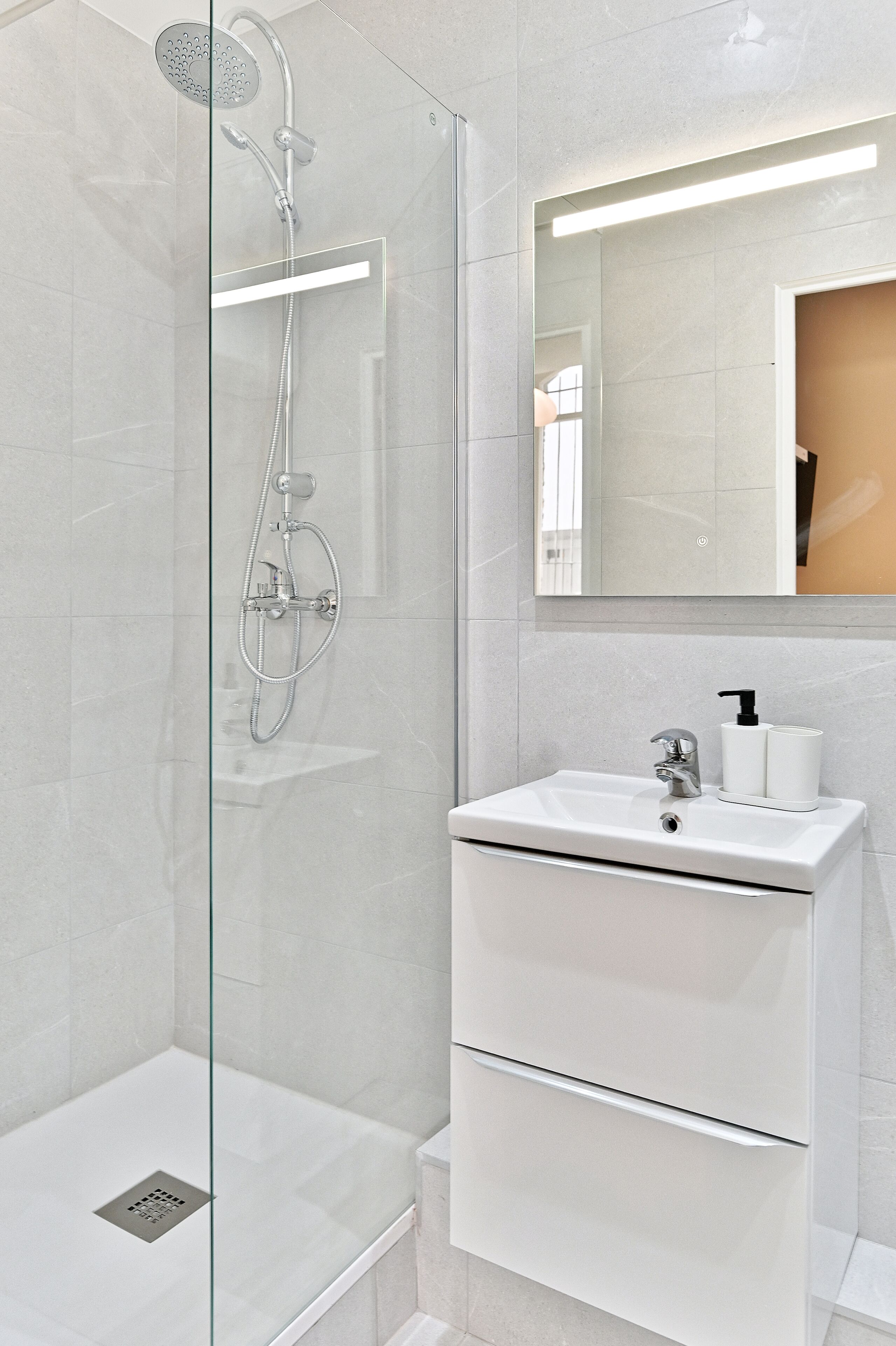 A modern bathroom with a glass-enclosed shower, a white sink with a soap dispenser, and a mirror above the sink.