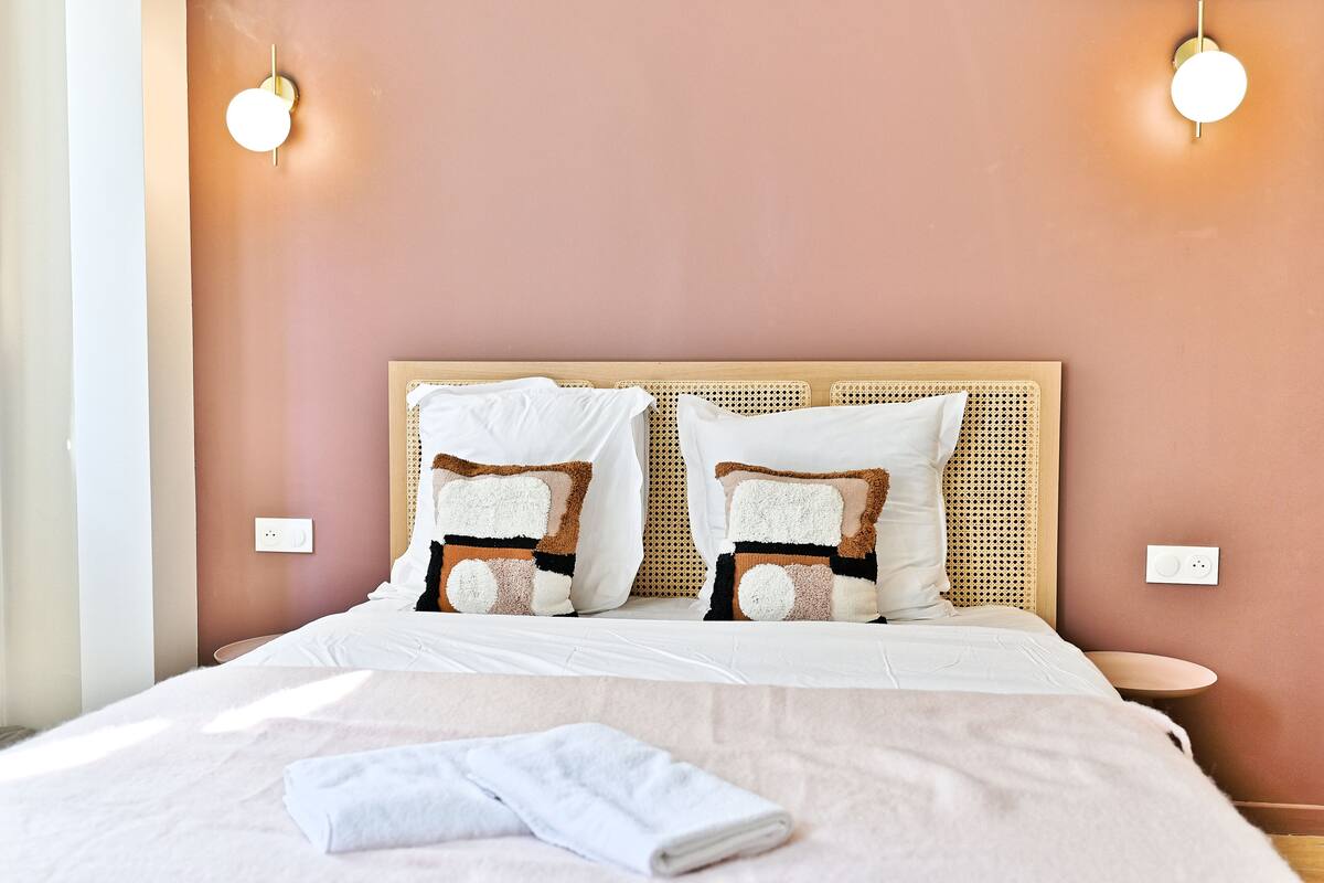 Un letto rifatto con lenzuola bianche e cuscini decorativi, un comodino rotondo e un lampadario fissato alla parete.