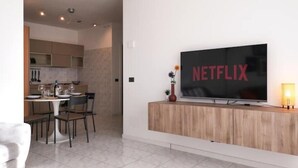 A modern living room with a large flat-screen TV mounted on a wooden console, a dining area with a table and chairs, and a kitchen in the background.