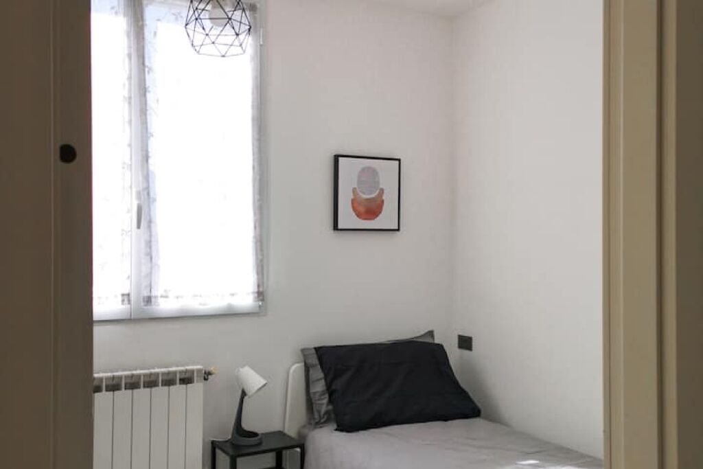 Una habitación pequeña y poco amueblada con una cama, un radiador, un escritorio con una lámpara y una ventana con una lámpara de diseño geométrico.