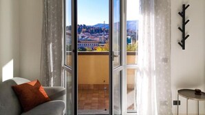 A room with a view of a cityscape through a glass door, a grey sofa with an orange pillow, and a small table with a candle.
